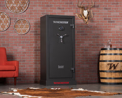 A Winchester Safe Bandit 19 Gun Safe in slate color stands against a brick wall, providing secure firearm storage near a red chair, a barrel, and decorative items.