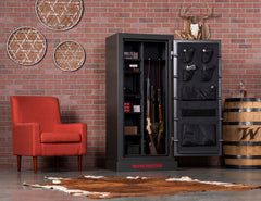 The Winchester Bandit 19 Gun Safe Slate by Winchester Safe is open in a room featuring a red chair, brick wall, decorative hoops, wooden barrel, and cowhide rug on the floor, showcasing secure firearm storage with style.