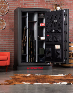 A Winchester Legacy 53 Fireproof Gun Safe by Winchester Safe stands open against a brick wall, showcasing rifles, handguns, and accessories. The door includes storage pouches using the GunStiXX system. Nearby are a red chair, barrel, animal hide rug, and circular wall decor.